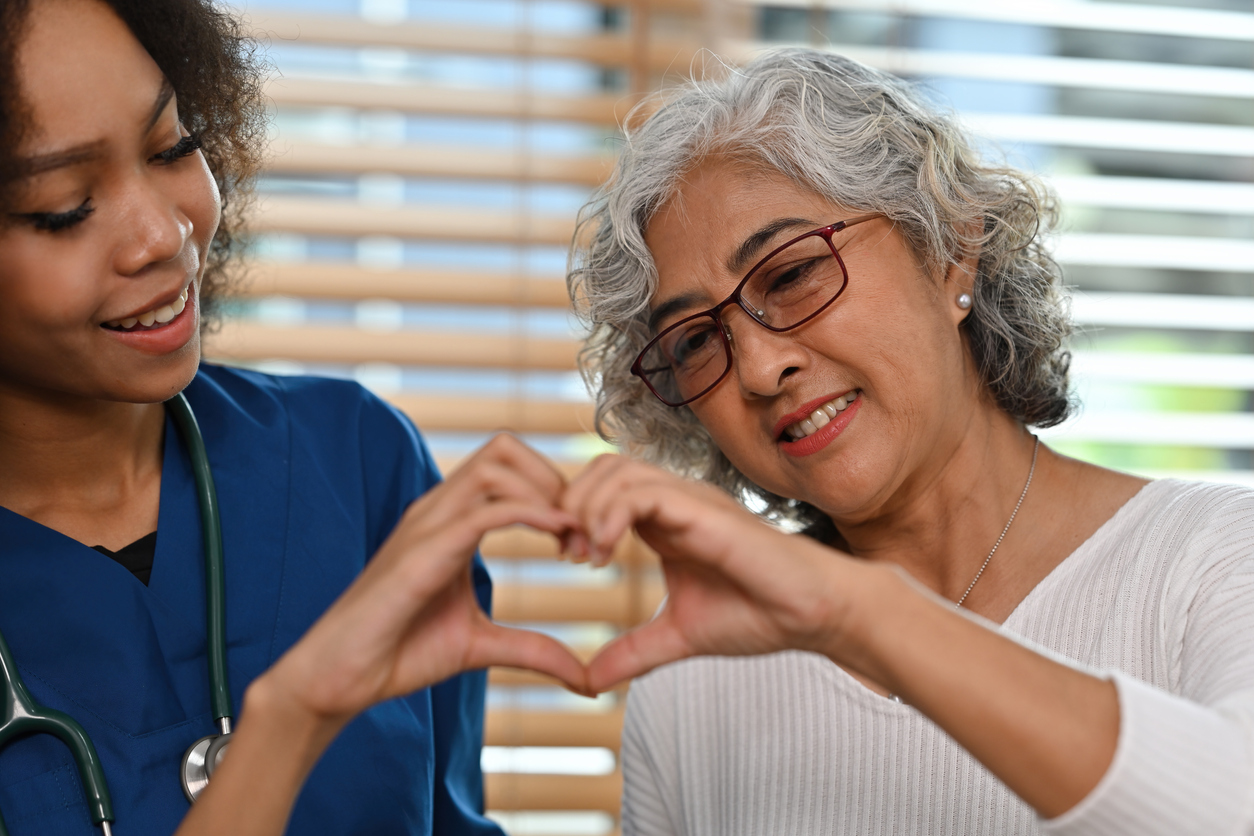A lovely elderly patient woman and African caregiver are making a heart shape together, holding hands symbolizing love and care, Caring for the elderly people and nursing home concepts